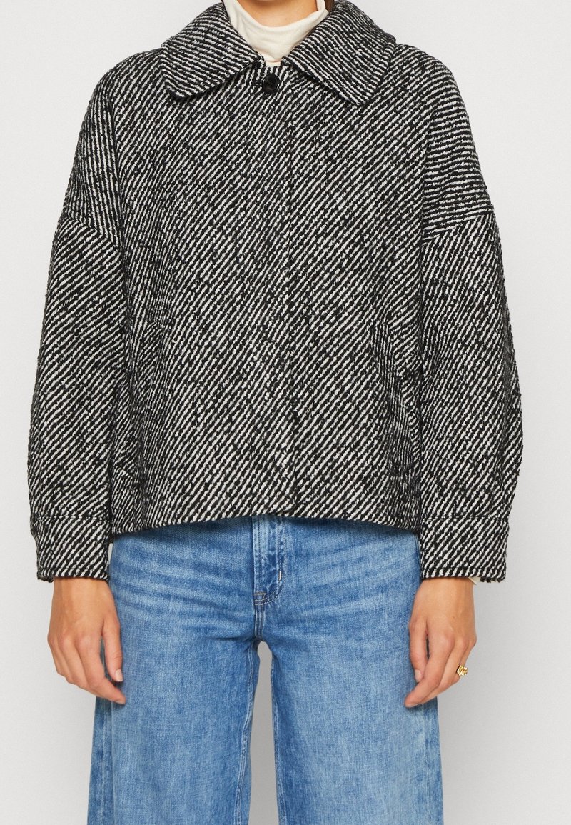 Strukturalna, czarno-biała kurtka o kroju nyższym, z szerokim kołnierzem i dużymi guzikami, połączona z jasnoniebieskimi spodniami w szerokim kroju.