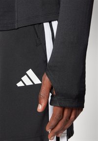 Schwarzes Sport-Shirt mit langen Ärmeln, Daumenlöchern und Reißverschlusstasche. Mit weißem Drei-Streifen-Design und auffälligem weißen Logo auf den Shorts.
