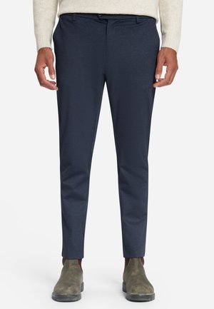 Mann trägt marineblaue, schmal geschnittene Hose, beigen Pullover und braune Leder-Stiefeletten und steht vor einem einfarbigen weißen Hintergrund.