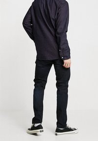 Camicia a quadri blu navy con maniche lunghe, abbinata a jeans scuri e sneakers nere, vista da dietro.