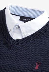 Laivastonsininen neulepusero, jonka alla valkoinen kauluspaita. Rinnassa on punainen hirvimerkki ja kankaan tekstuuri on kuvioitu.