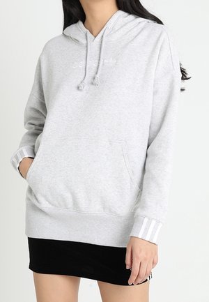 Vrouw draagt een lichtgrijze Adidas hoodie met voorvak en witte strepen op de boorden, gecombineerd met een zwarte rok.