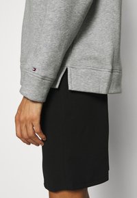 Man med en grå långärmad sweatshirt med en liten logotyp nära manschetten och svarta shorts, visad från sidan på midjan.