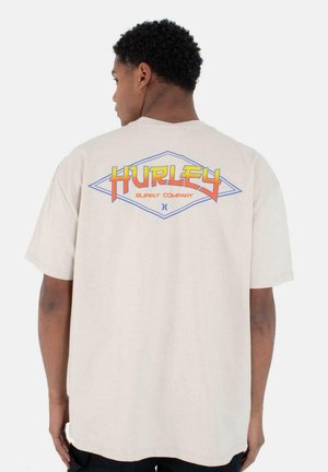 Camiseta de manga corta color beige con un colorido gráfico de "Hurley Supply Company" en la parte posterior dentro de una forma de diamante. Textura de algodón suave.