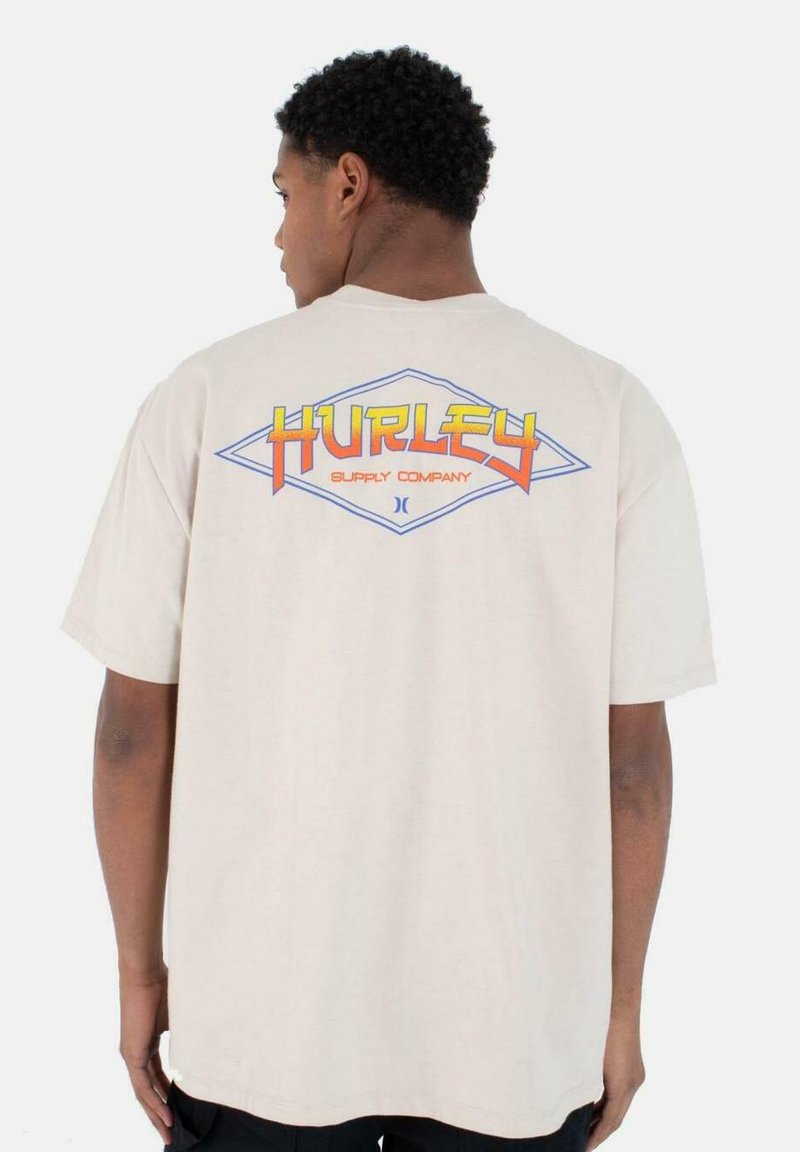 Camiseta de manga corta color beige con un colorido gráfico de "Hurley Supply Company" en la parte posterior dentro de una forma de diamante. Textura de algodón suave.
