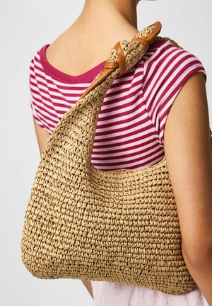 Femeie care poartă o cămașă în dungi roșii și albe și care poartă o geantă mare de umăr împletită din paie cu o curea înnodată.