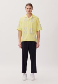 Camicetta gialla a maniche corte con stampe geometriche, vestibilità comoda, chiusura davanti con bottoni, abbinata a pantaloni scuri e sneakers bianche.