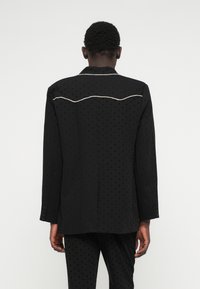 Czarna marynarka z fakturowanym wzorem w kropki, z falistym akcentem z perełek na górnej części pleców i zapięciem na guziki przy mankietach.