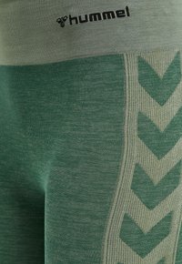 Detailní záběr na zeleno-šedé sportovní legíny s logem Hummel a chevronovým vzorem na bočním materiálu.