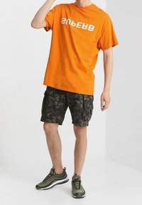 T-shirt in cotone arancione con testo bianco, abbinato a pantaloni cargo mimetici scuri e sneakers verde oliva. Look casual, vestibilità rilassata.
