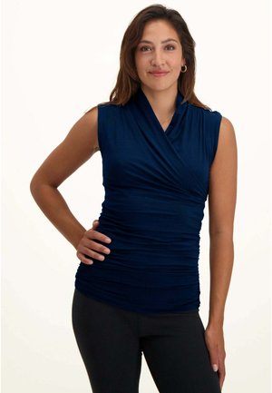 Femme aux cheveux châtains mi-longs portant un haut bleu marine sans manches froncé et un legging noir, souriante avec une main sur la hanche.