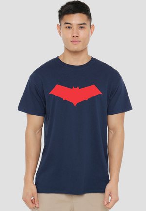 Man wearing navy blue t-shirt with large red stylized bat symbol on chest, standing against plain light gray background.
