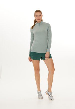 Hellgrünes Langarmhemd mit einem halben Reißverschluss, kombiniert mit dunkelgrünen Shorts. Das Model trägt graue und schwarze Sportschuhe. Glattes Material.