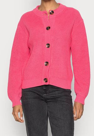 Cárdigan tejido de color rosa brillante con cinco grandes botones oscuros, llevado sobre unos vaqueros negros por una persona que está de pie frente a un fondo liso.