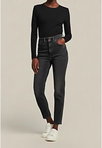 Femme portant un haut à manches longues noir, un jean taille haute foncé et des baskets blanches, debout avec une main dans la poche.