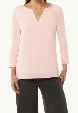 Kvinde iført en lyserød langærmet bluse med rund, delt halsudskæring og mørkegrå bukser, stående med hænder afslappet ned langs siden.