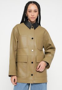 Kaķa ādas jaka ar melnu apkakli, divām lielām priekšējām kabatām, aizsprādzējamiem pogu aizdariem un redzamām šuvju detaļām.