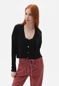 Schwarze gerippte Strickjacke mit V-Ausschnitt, versehen mit goldenen Knöpfen. Kombiniert mit staubrosa Stoffhosen mit Taillenknoten, die eine lockere Passform betonen.