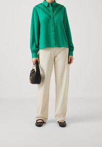 Camicia a maniche lunghe verde con dettagli in pizzo, abbinata a pantaloni a gamba larga bianco sporco e borsa nera. Ballerine nere con decorazioni.