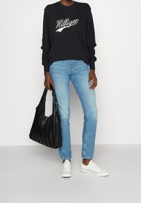 Sudadera negra con logo blanco de "Hilfiger", vaqueros de denim azul claros y zapatillas blancas. Bolsa negra de pliegues con textura suave, sostenida con una mano.