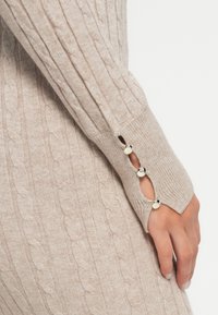 Manga de punto beige con tres botones plateados cerca de la muñeca, cubriendo parcialmente una mano con uñas naturales.