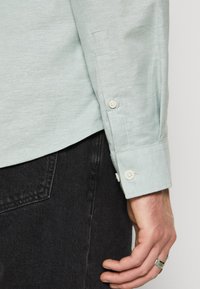 Gros plan sur la main d'une personne portant une bague en argent, un poignet de chemise à manches longues vert clair avec des boutons et une poche de pantalon en denim noir.