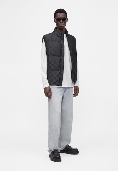 Homme portant une doudoune noire à carreaux, une chemise blanche à manches longues, un jean bleu clair à jambes larges, des chaussures noires et des lunettes de soleil foncées sur fond blanc.