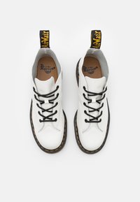 Zapatos de cuero blancos con cordones negros, puntera redonda y suela de goma negra. Costuras amarillas a lo largo del borde. Tirador negro con logotipo.