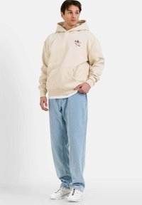 Beige hoodie med ficka framtill och broderad design. Kombination med ljusblå jeans och vita sneakers. Modellen står mot en enfärgad bakgrund.