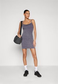 Violet besiūlis suknelė su prie kūno prigulančiu dizainu, pagaminta iš minkštos medžiagos, derinama su juoda rankine ir juodais sportiniais bateliais, stovinti ant vienodos fono.