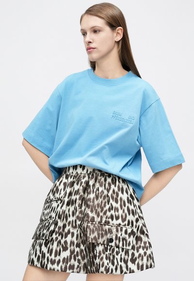 Jeune femme portant un t-shirt bleu oversize avec un texte et une jupe taille haute à imprimé léopard avec de grandes poches, posant devant un fond uni.