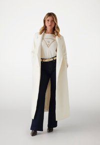 Largo abrigo blanco hecho de una tela suave, combinado con una blusa crema que presenta un adorno triangular, jeans acampanados oscuros y zapatos de tacón.