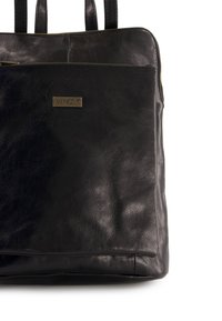 Czarna skórzana torba typu tote o sztywnym kształcie, z metalową plakietką w kolorze złota, zamek błyskawiczny i podwójne uchwyty. Gładka faktura.