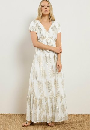 Donna che indossa un abito lungo bianco con motivi floreali dorati, maniche corte, scollo a V e vita arricciata, in piedi su un pavimento di stuoia intrecciata.