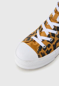 Καμβά παπούτσι τύπου sneaker με λεοπάρ μοτίβο σε κίτρινο και μαύρο, λευκή λαστιχένια μύτη και λευκά κορδόνια. Υφασμάτινη υφή με μεταλλικές τρύπες.