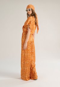 Tuta arancione a fantasia con maniche corte, vita alta legata e foulard coordinato. Modella indossa occhiali da sole. Tessuto morbido e vestibilità ampia.