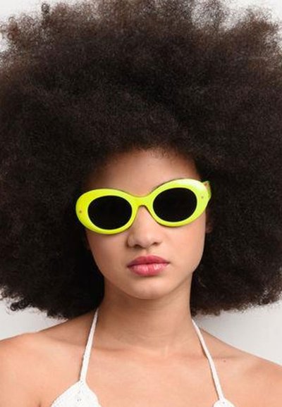 Occhiali da sole giallo neon con un design elegante e arrotondato e lenti scure. Indossati con un top bianco a collo alto e circondati da folti riccioli scuri.