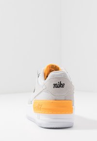 Nike tenisky se světle šedým svrškem, oranžovým detailem na patě, bílou mezipodešví, texturou semiše a logem na zadní straně.