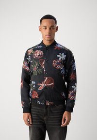 Μαύρο bomber jacket με λουλούδια και σχέδια σε κόκκινο, μπλε και πράσινο. Διαθέτει κουμπωτή μπροστά και ριμπ μανσέτες. Φορεμένο πάνω από σκούρα τζιν.