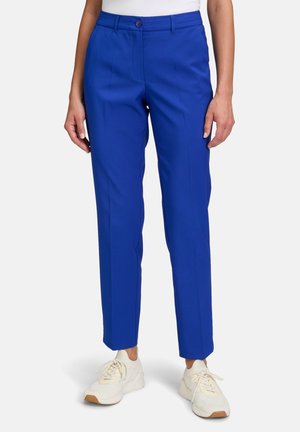 Pantalon ajusté bleu avec une texture lisse, comprenant une fermeture à un seul bouton, des plis avant et des jambes fuselées, associé à des baskets blanches.
