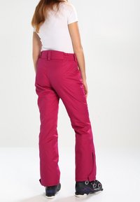 Fuchsia skibroek met een verstelbare tailleband, rechte pijpen en ritsen aan de zoom, gecombineerd met zwarte skischoenen.