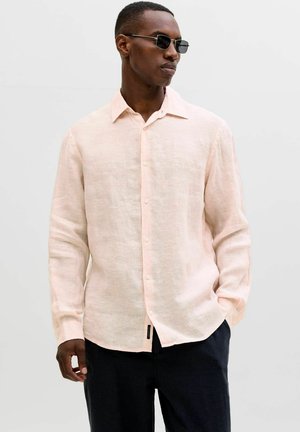 Homme portant une chemise boutonnée à manches longues de couleur rose clair, un pantalon foncé et des lunettes de soleil rectangulaires, debout avec une main dans la poche.