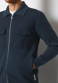 Marinblå zip-up jacka med texturerade stickade paneler och släta tygsektioner. Har två framfickor med klaffstängningar och sidofickor.
