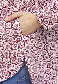 Chemise à motifs en tissu blanc avec des motifs circulaires rouges, une poche latérale et un ourlet arrondi. Main visible dans la poche.