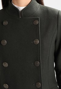 Tmavě zelený vlněný kab�át s mandarínkovým límcem a šesti ozdobnými kovovými knoflíky s květinovým vzorem na přední straně.