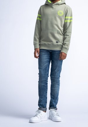 Bluza z kapturem w oliwkowym kolorze z bawełny, z odblaskowymi żółtymi paskami i logo, w połączeniu z dopasowanymi niebieskimi dżinsami i białymi trampkami.