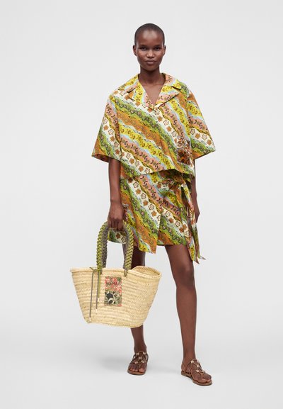 Femme portant un ensemble chemise et jupe à rayures colorées, tenant un grand sac en paille tressée et portant des sandales marron, debout sur fond blanc.