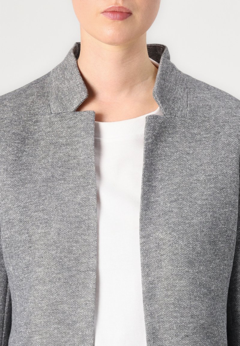Graue strukturierte Jacke mit einer strukturierten Oberfläche und einem Stehkragen, getragen über einem weißen Oberteil. Klare Linien und minimalistisches Design.