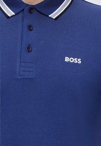 Modré polo tričko s bielym a čiernym pruhovaným golierom, tromi tmavými gombíkmi a bielym logom "BOSS" na ľavej strane hrudníka.