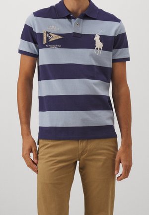 Homme portant un polo rayé bleu marine et bleu clair avec des logos de yacht et de joueur de polo, associé à un pantalon beige, sur un fond uni.